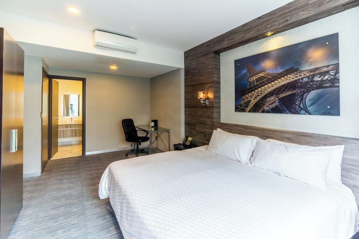 Quarto de hotel com uma cama grande, uma escrivaninha com cadeira, um quadro da Torre Eiffel e um ar condicionado.