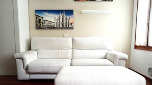 A living room with a white sofa and ottoman, a painting of a cathedral, and a window with curtains. - Lovely Flat - Magolfa 21 (Milan)