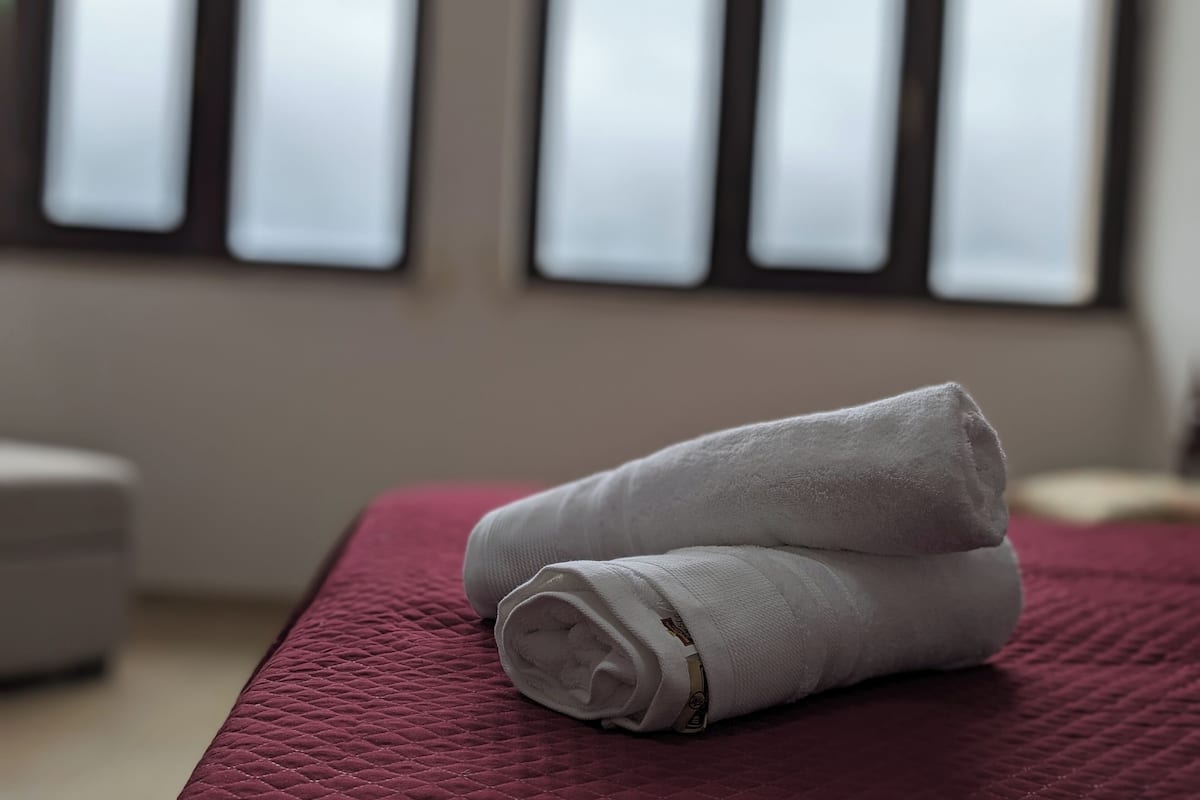 A neatly made bed with a red quilted bedspread and two rolled white towels on it.