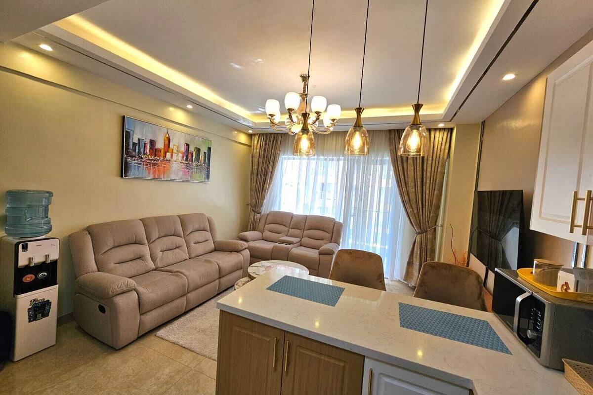 A modern living room with a sofa set, a dining table, and a kitchen area with a refrigerator and microwave.