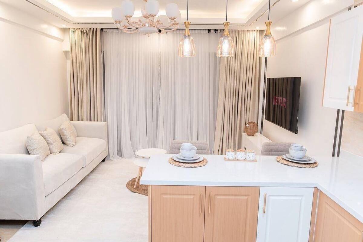 A modern living room with a white sofa, a kitchen area with a white countertop, and a large window with curtains.