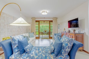 Uma sala de estar com sofá, televisão, área de jantar e vista através de uma porta de vidro.