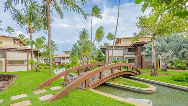 Een resort met een houten brug over een waterpartij, omgeven door weelderige begroeiing en palmbomen.