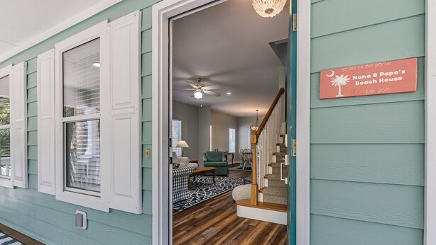 A wooden staircase leading to a living room with a ceiling fan, a sign reading "Nana & Papa's Beach House," and a view of the outside. - Golf cart included, amenity cards available, pet friendly - great for young families, 601 Porpoise Circle (Fripp Island)