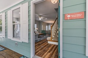 A wooden staircase leading to a living room with a ceiling fan, a sign reading "Nana & Papa's Beach House," and a view of the outside.