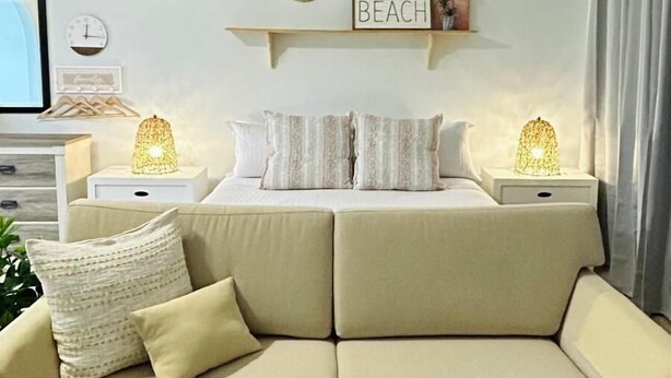Un salon avec un canapé beige, deux tables de chevet avec des lampes, une horloge murale et un panneau indiquant « PLAGE ».