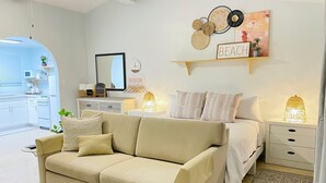 Une chambre moderne avec un canapé beige, un lit avec une literie blanche, une table de chevet avec une lampe, une étagère murale agrémentée d’objets décoratifs et une vue sur la cuisine.