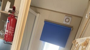A compact bathroom with a toilet, a window with a blue blind, and a fire extinguisher mounted on the wall. - Coastal Cabin Retreat in Rhyl (Rhyl)