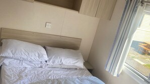 A bed with white bedding and a headboard, a window with curtains, and a wall-mounted shelf unit. - Coastal Cabin Retreat in Rhyl (Rhyl)