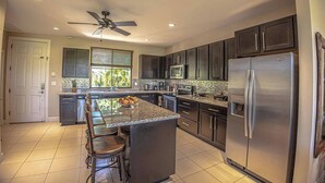 Una cocina moderna con gabinetes oscuros, electrodomésticos de acero inoxidable, una isla central con taburetes y un ventilador de techo.