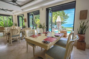 Un espace repas offrant une vue sur l’océan, comprenant une grande table avec des chaises, un ventilateur de plafond et des plantes en pot.