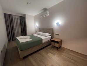 Ein ordentlich bezogenes Bett mit grüner Tagesdecke und weißen Kissen, ein Nachttisch aus Holz mit Schublade, eine Wand-Klimaanlage und ein Fenster mit Vorhängen.