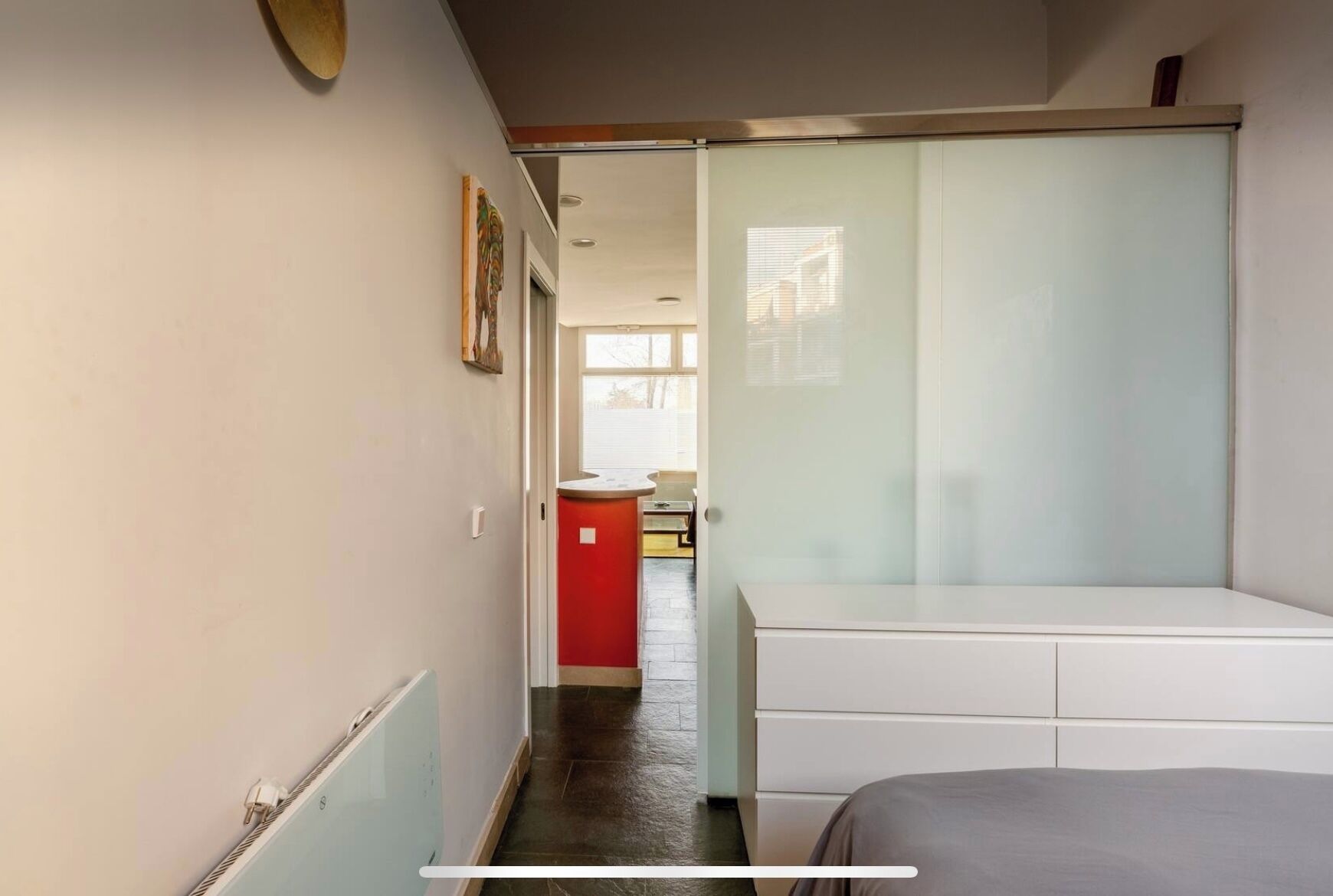 Un pasillo estrecho que conduce a una habitación con una cama, un armario rojo y una ventana con persianas.