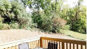 A deck with a glass table and chairs, surrounded by trees and a dirt path.