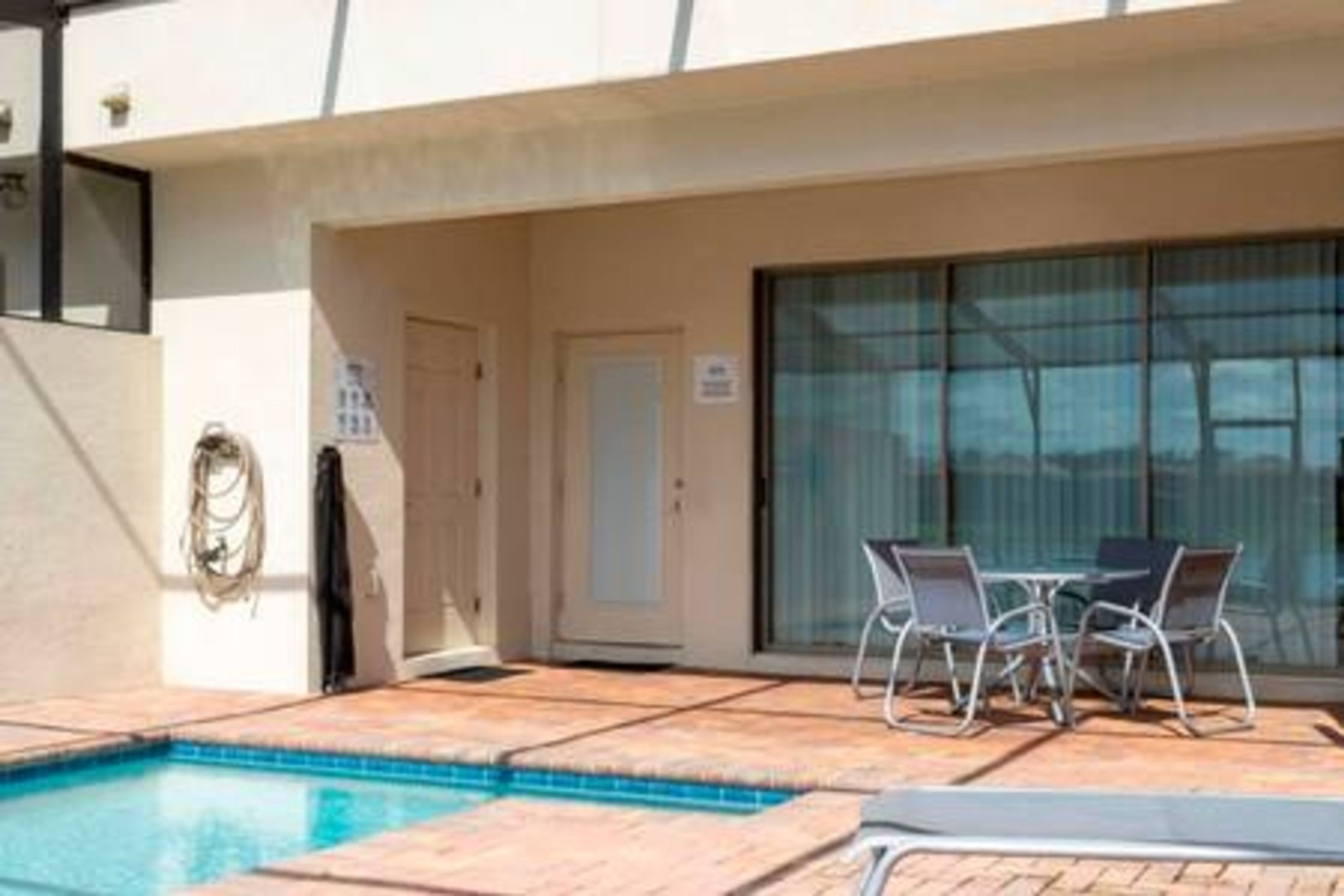Area piscina con solarium piastrellato, un tavolo e sedie, e una porta scorrevole in vetro.