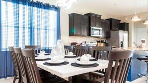 Un comedor con una mesa dispuesta para cuatro, sillas de madera oscura y una cocina al fondo con armarios y electrodomésticos oscuros.