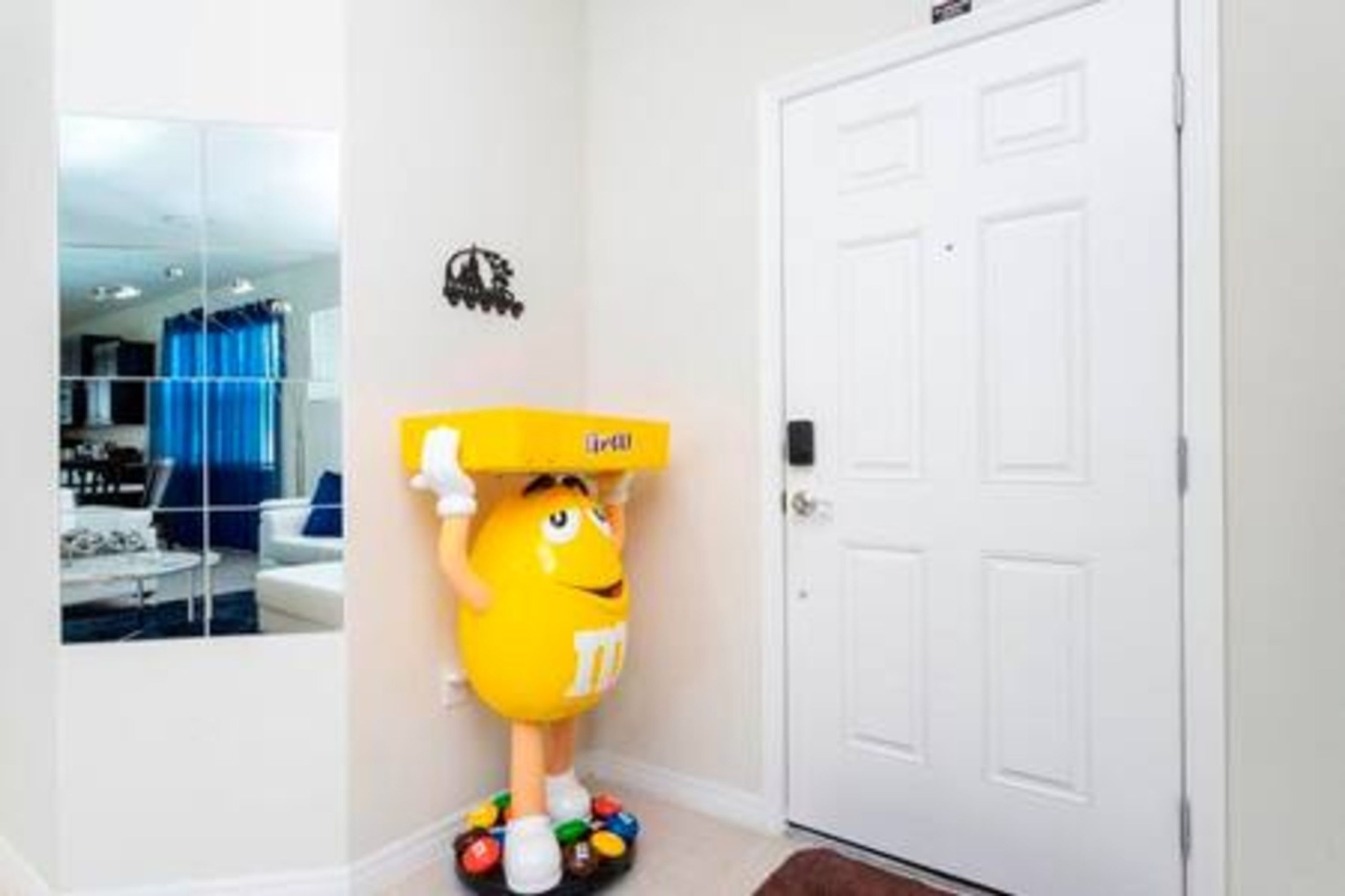 A room with a white door, a mirror reflecting a blue curtain, and a yellow object resembling a character with a box on its head.