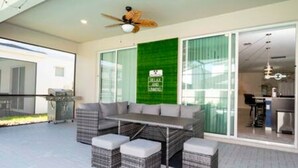Un espacio exterior moderno con área de descanso, un sofá y otomanas, un ventilador de techo y una pared verde con un letrero.