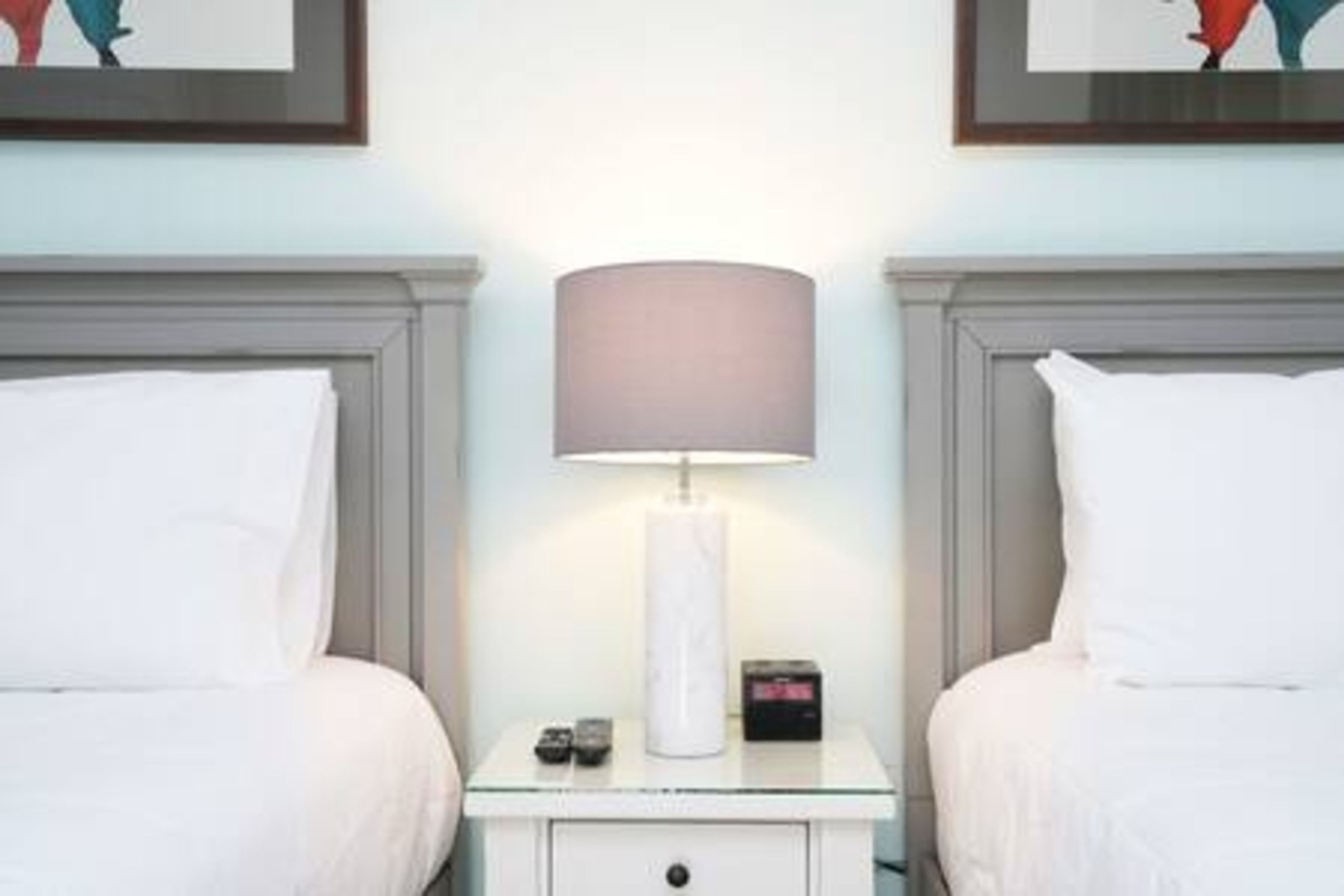 Camera d'albergo con due letti, un comodino con una lampada, una sveglia e un telecomando.