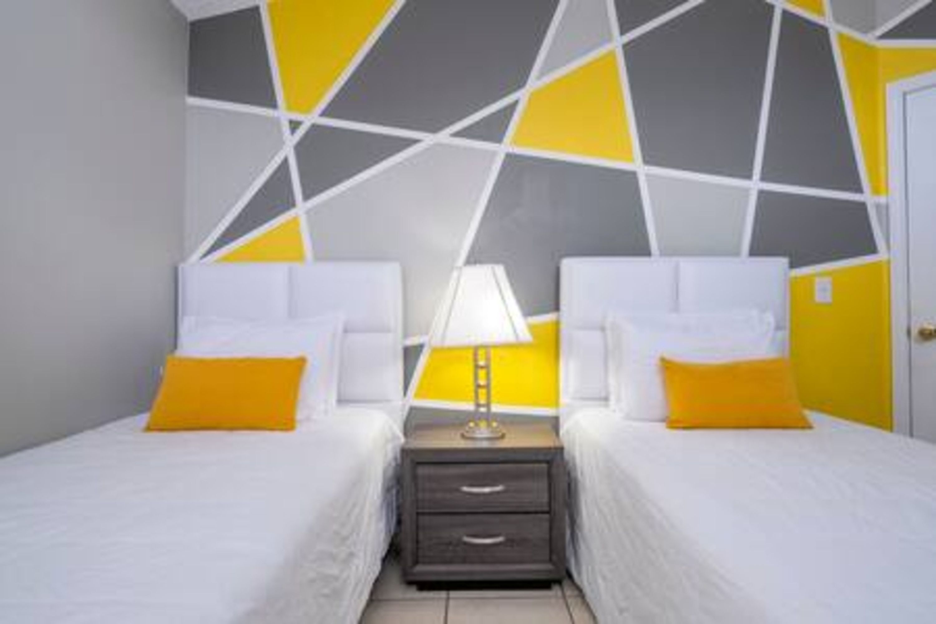 Habitación de hotel con dos camas, una mesita de noche con lámpara y un muro con diseño geométrico.