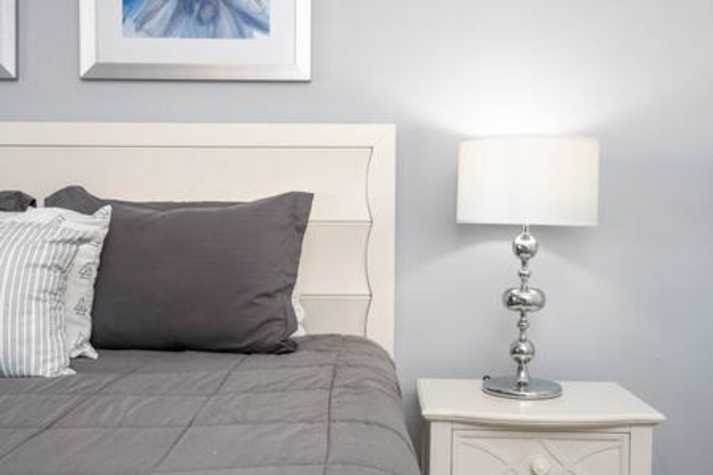Una cama bien tendida con edredón y almohadas grises, una mesita de noche con una lámpara y un cuadro enmarcado en la pared.