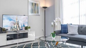 A modern living room with a flat-screen TV, a glass coffee table, a grey sofa, and a decorative plant. - Gorgeous Home With Pool and Lake View (Kissimmee)