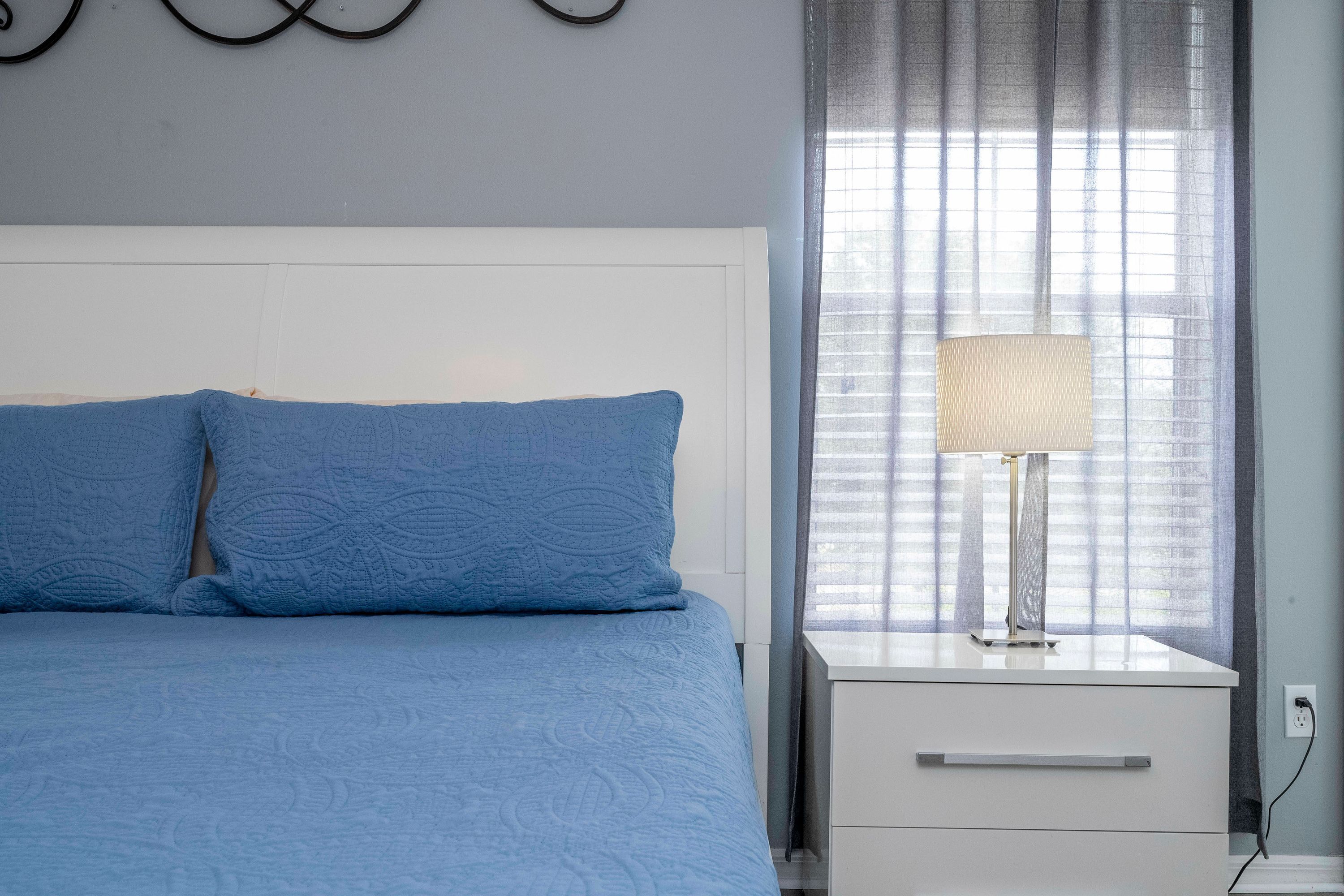 Un dormitorio con cama, mesita de noche, lámpara y ventana con cortinas.