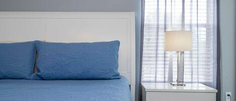 Un dormitorio con cama, mesita de noche, lámpara y ventana con cortinas.