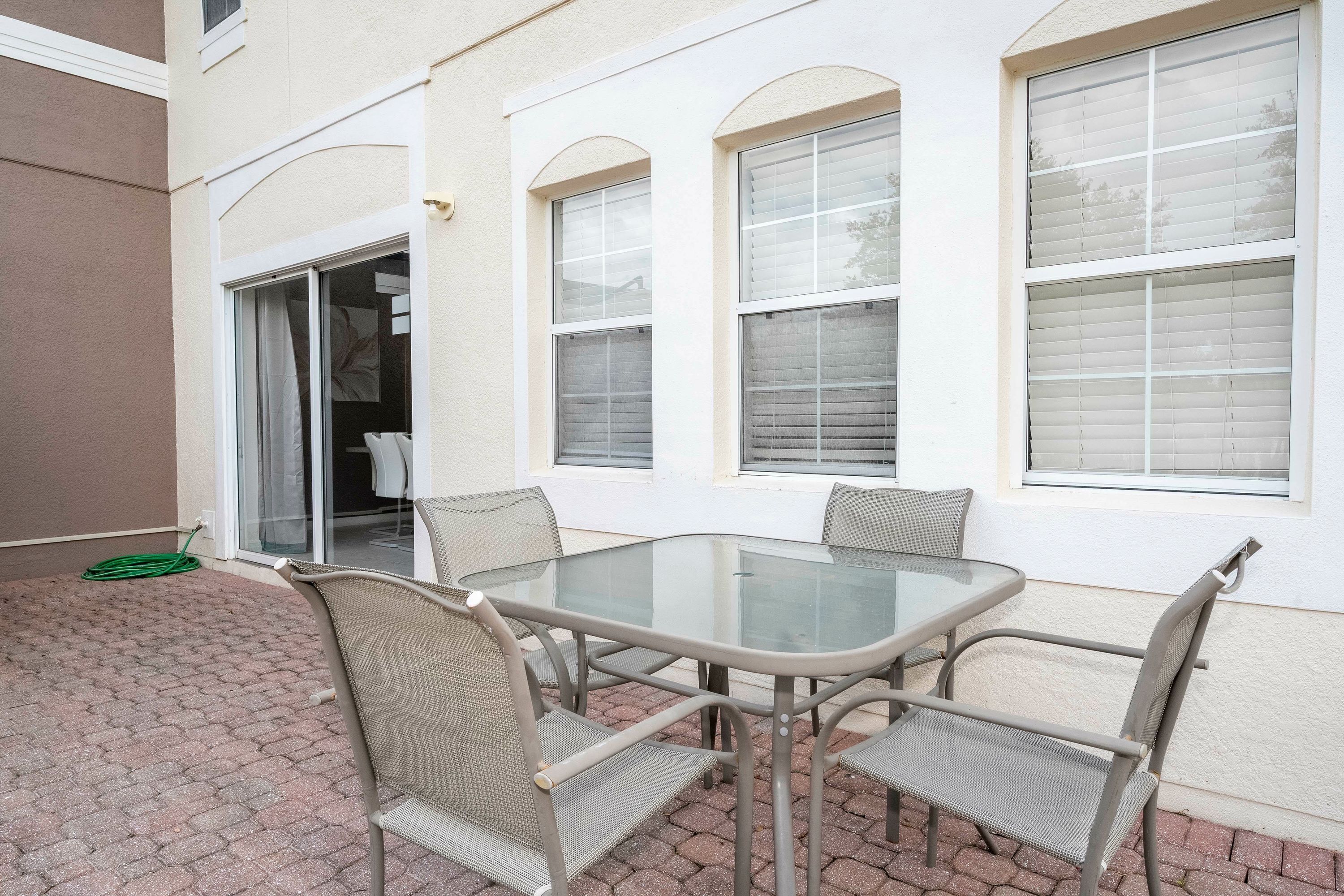 Une terrasse extérieure avec une table en verre et quatre chaises, un tuyau d’arrosage vert et une porte-fenêtre coulissante donnant sur un espace abrité.