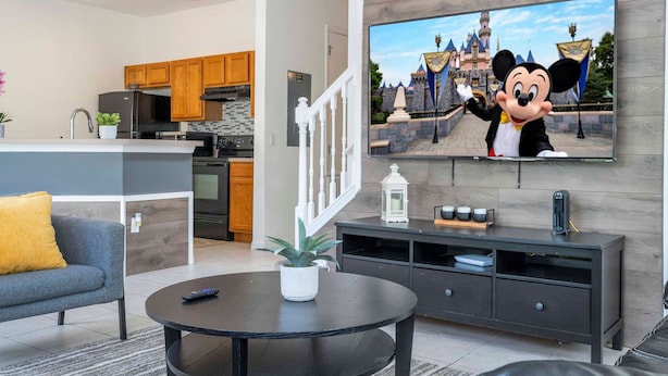 A modern living room with a flat-screen TV displaying a cartoon character, a dining area with wooden cabinets, and a kitchen with stainless steel appliances. - 4 Bedrooms Close to Disney (Kissimmee)