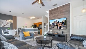 A modern living room with a black leather sofa, a gray armchair, and a coffee table. There is a TV mounted on the wall displaying an animated character. The room has a wooden floor and a ceiling fan. - 4 Bedrooms Close to Disney (Kissimmee)