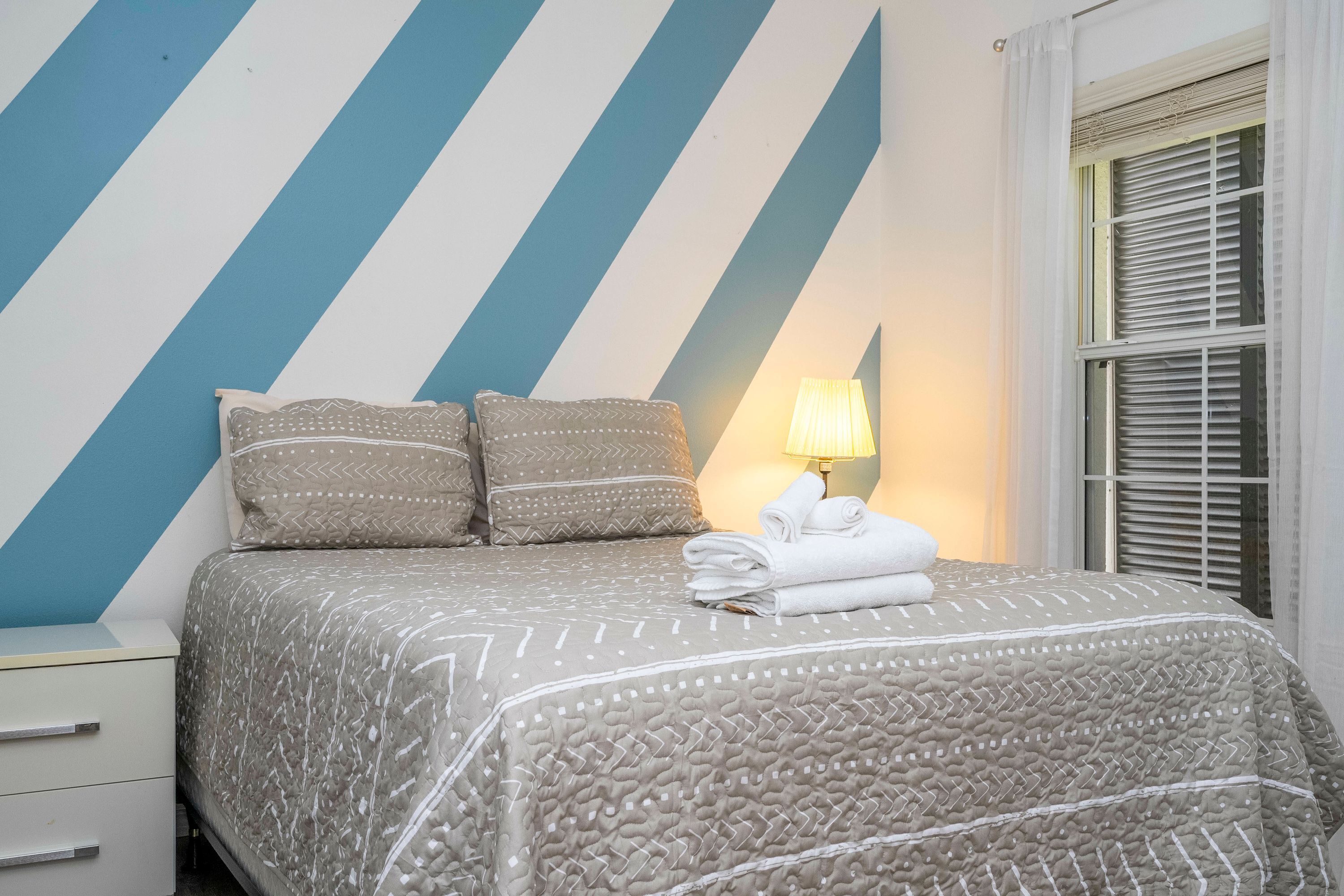 A neatly made bed with patterned bedding, a bedside lamp, and a window with blinds.