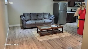 Een woonkamer met een grijze bank, een houten salontafel en een keuken met donkere kasten en apparatuur.