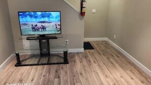 Een kamer met een flatscreen televisie op een glazen voetstuk, een brandblusser aan de muur en houten vloeren.
