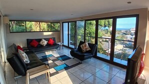 A spacious living room with a sofa, armchairs, and a coffee table. Large windows offer a view of a cityscape and greenery. - Cozy Margate Retreat Family Home With 2 Bedrooms (Margate)