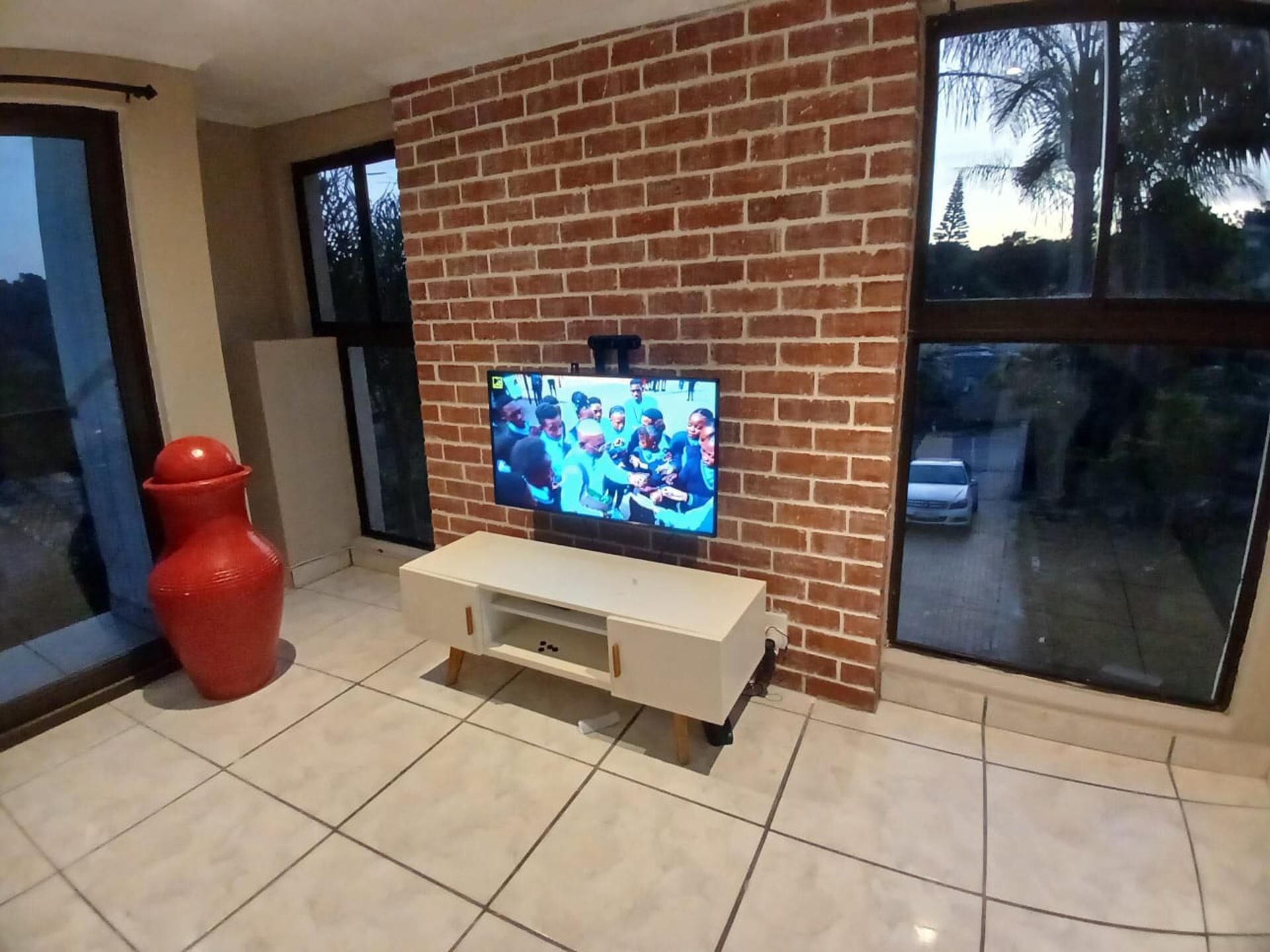 A living room with a television on a brick wall, a white entertainment unit, a red vase, and large windows with a view of palm trees.