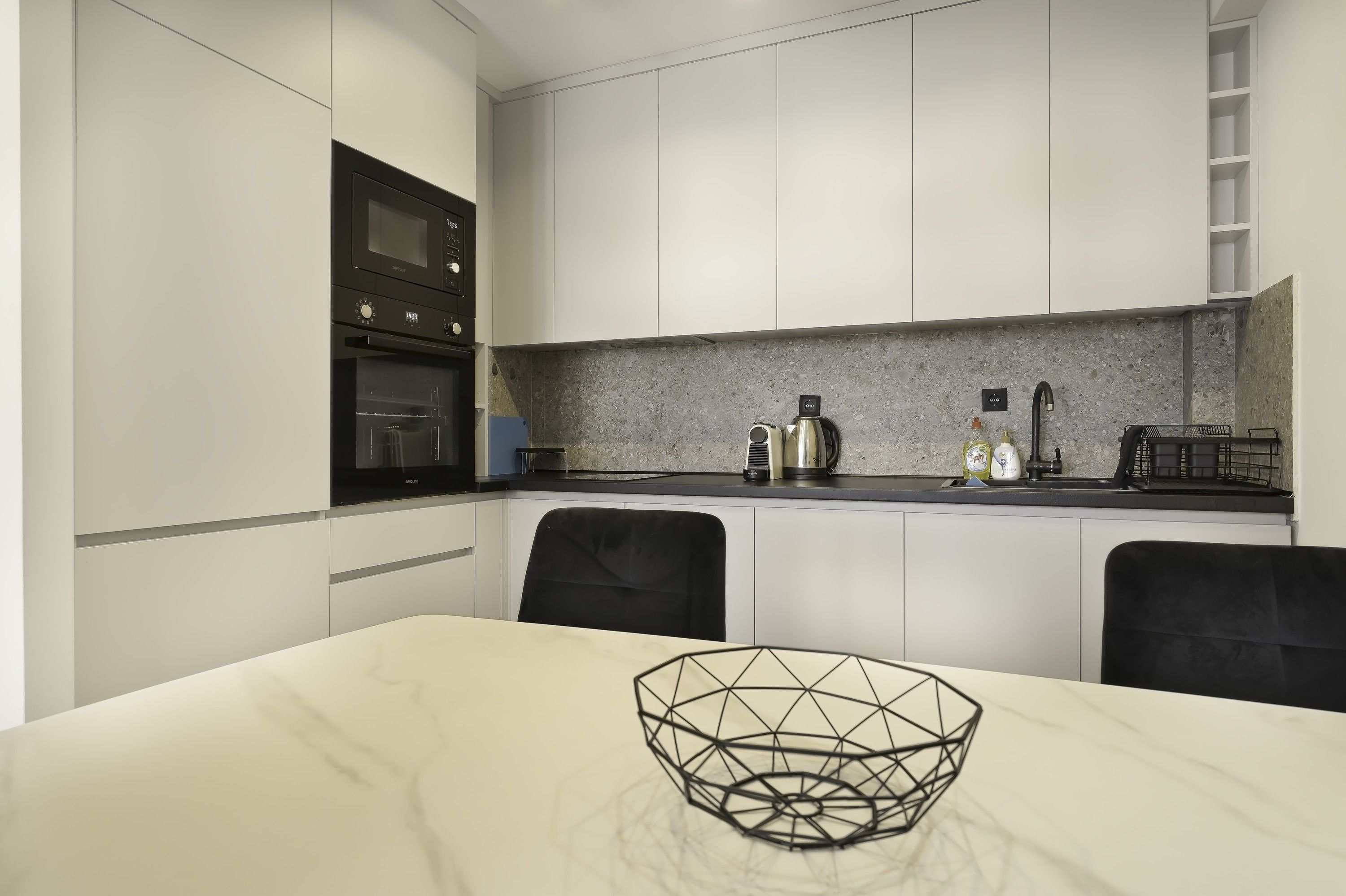 A modern kitchen with white cabinetry, a built-in oven, a coffee maker, and a kettle on the countertop.