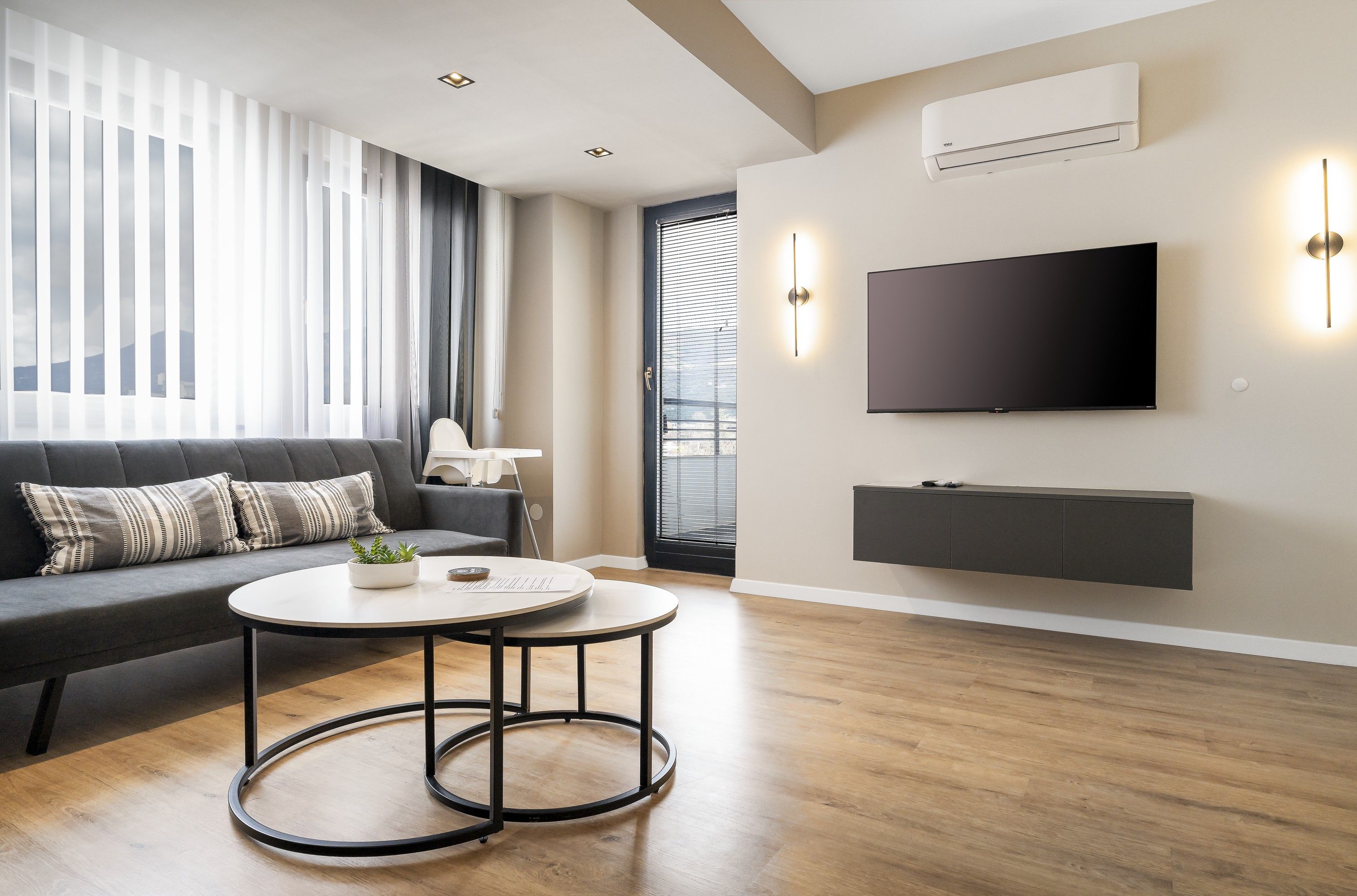 A modern living room with a sofa, a round coffee table, a flat-screen TV mounted on the wall, and a built-in shelf.