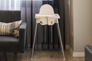 A high chair with a tray table, positioned next to a dark-colored sofa with a patterned pillow.