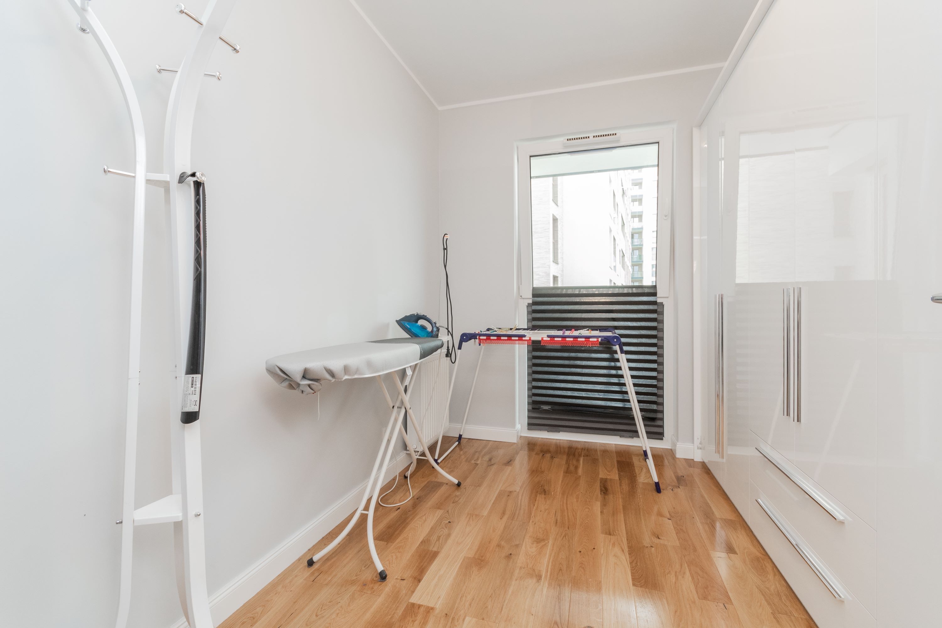 Une pièce de taille modeste, avec un plancher en bois, un radiateur blanc, une fenêtre donnant sur des immeubles et un étendoir à linge.