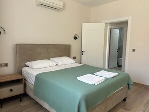 A neatly made bed with white pillows and a green bedspread, a wooden nightstand, and a white door leading to a bathroom. - White Suites Icmeler (Içmeler)