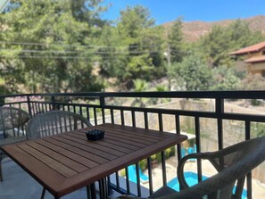 A wooden table with a black object on it, a wicker chair, and a view of a swimming pool and trees. - White Suites Icmeler (Içmeler)