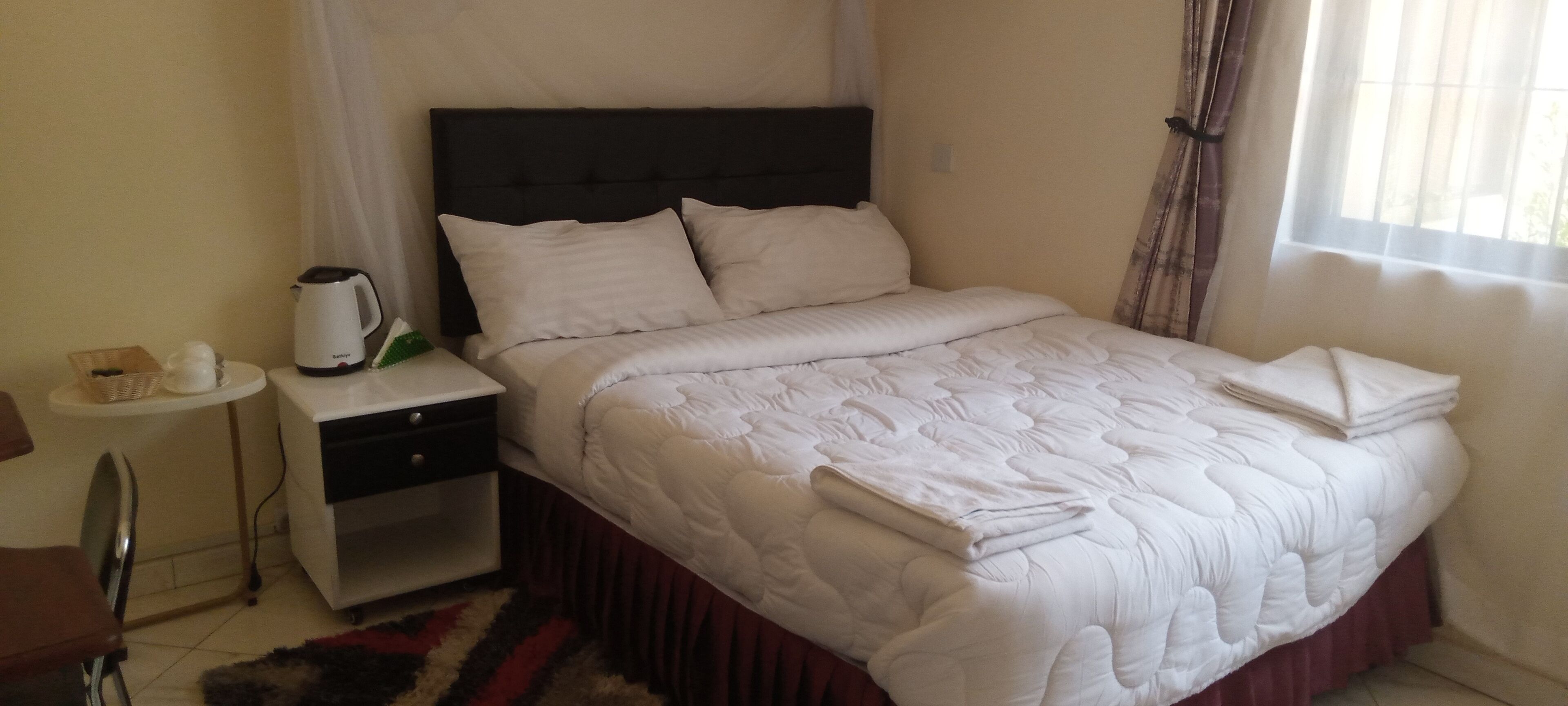 Una cama bien hecha con un edredón blanco y almohadas, una mesita de noche con una tetera y una caja de pañuelos, una silla y una ventana con cortinas.