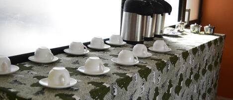 Una mesa con varias tazas y platillos blancos, un termo de acero inoxidable y pequeños recipientes, probablemente para té o café, frente a una ventana con vistas.