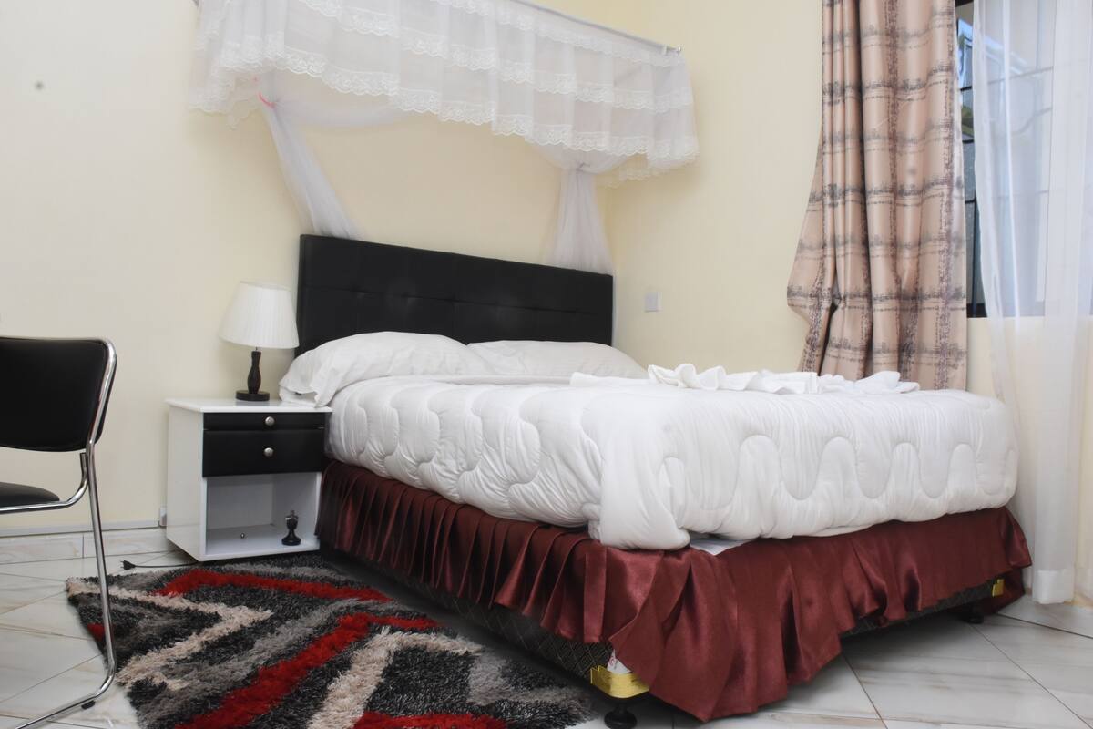 A bedroom with a bed, a nightstand, a chair, a bedside table, and a window with curtains.