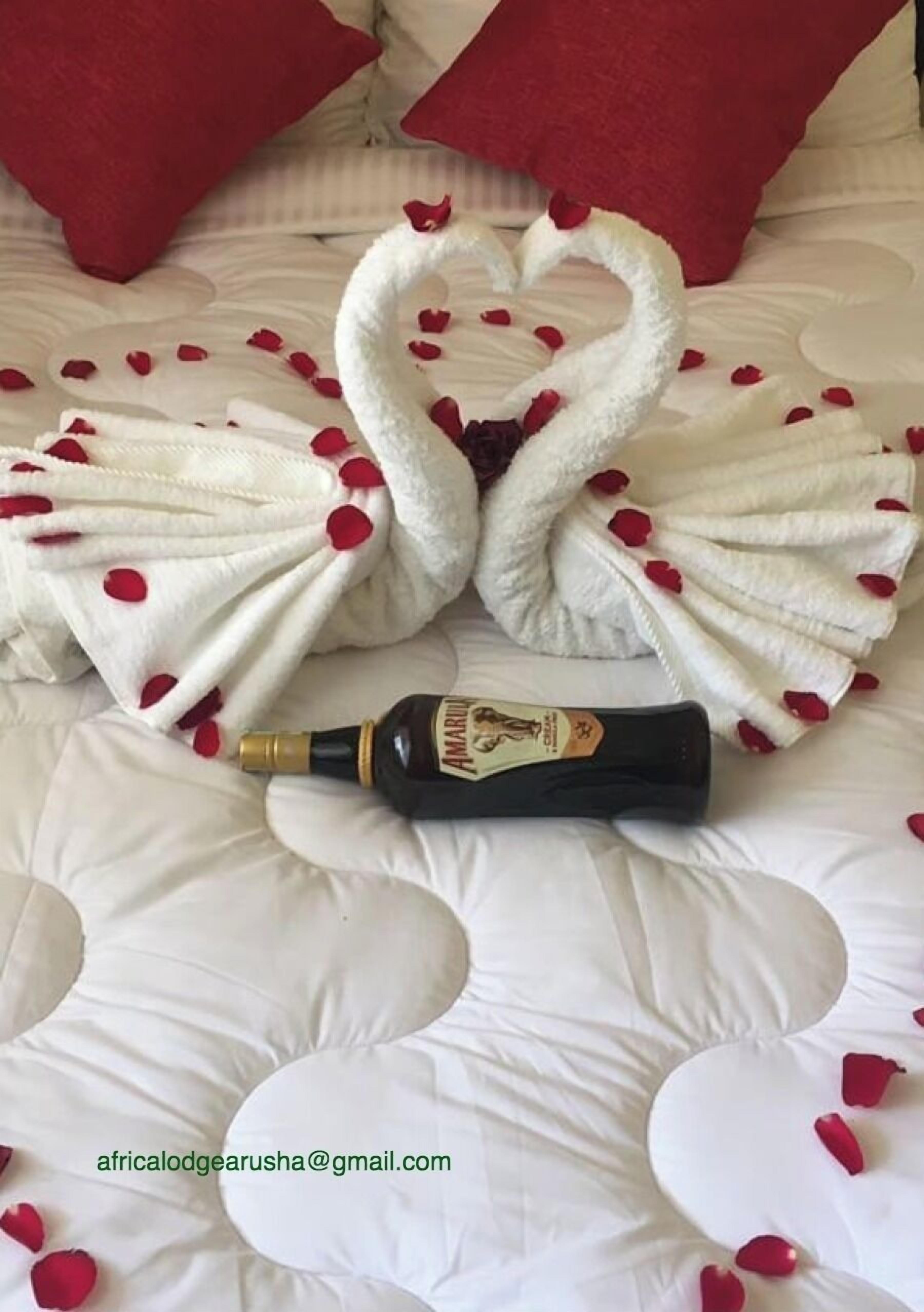 Un llit amb un edredó blanc, almoades vermelles, una ampolla d'Amaretto i una tovallola disposada en forma de cor amb pètals de rosa.