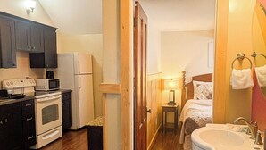 Cocina compacta con microondas, estufa y refrigerador, una mesita de noche pequeña con lámpara, y un baño con lavamanos y espejo.