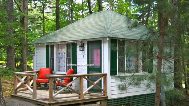Une petite cabane avec un toit vert et des murs blancs, dotée d’un porche avec une balustrade en bois et d’une chaise rouge.