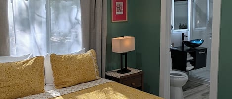 A bedroom with a bed, a bedside table with a lamp, a framed picture on the wall, and a view into the bathroom.