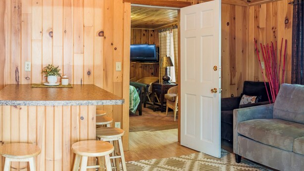 Ein Interieur einer Holzhütte mit Küchenzeile, Barhocker, einer Couch und einem Fernseher im Hintergrund.